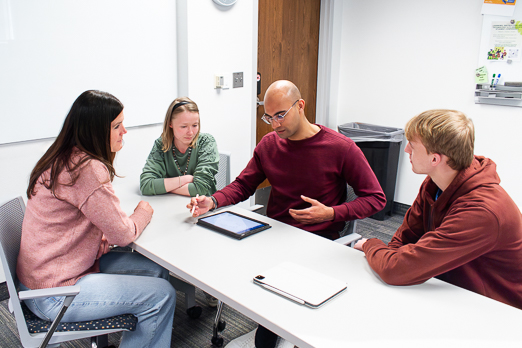 two men and two women at a long table while one man explains something on a tablet